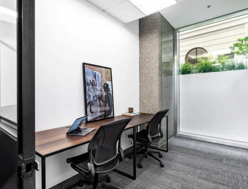 Two black mesh office chairs sit at a long wooden desk equipped with an open laptop, nestled in a minimalist workspace beside a textured column and frosted window. Text on the framed poster reads: "FLORENCE GUILD PRESENTS SIP & SKETCH. Art is size, style, a glass of wine and dynamic texture present in our life by showing your feelings, emotions and thoughts. Together, let's learn how to create art using charcoal, pencil, pastels and ink. Develop your drawing skills and unlock your inner creative power in this drawing session, whether you're a beginner or regular artist, everyone is welcome at this event. WORK CLUB LOCOMOTIVE TUESDAY, 7 MARCH 2024 5PM - 7PM"