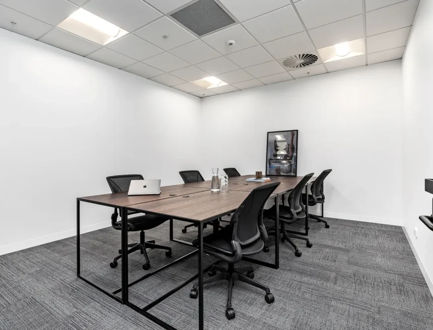 Black mesh chairs surround a long wooden conference table equipped with a laptop and water pitcher inside a bright, white-walled meeting room with grey patterned carpeting. Poster text reads: "NOW LIVE TRANSFORM YOUR HEALTH THE WAY YOU WORK"