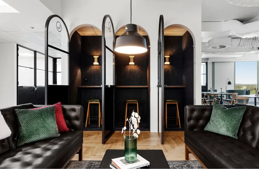 Three dark, arched phone booths equipped with stools stand side-by-side in a modern office interior behind two black leather couches and a coffee table featuring a book titled “ELEMENTS OF STYLE”.