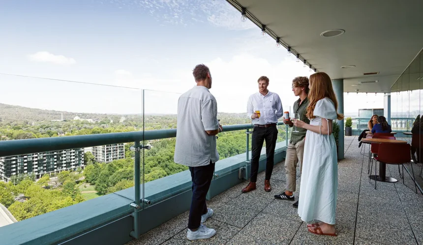 A group of four young adults chat while holding drinks on a covered high-rise balcony, overlooking a lush green city skyline through a glass railing.