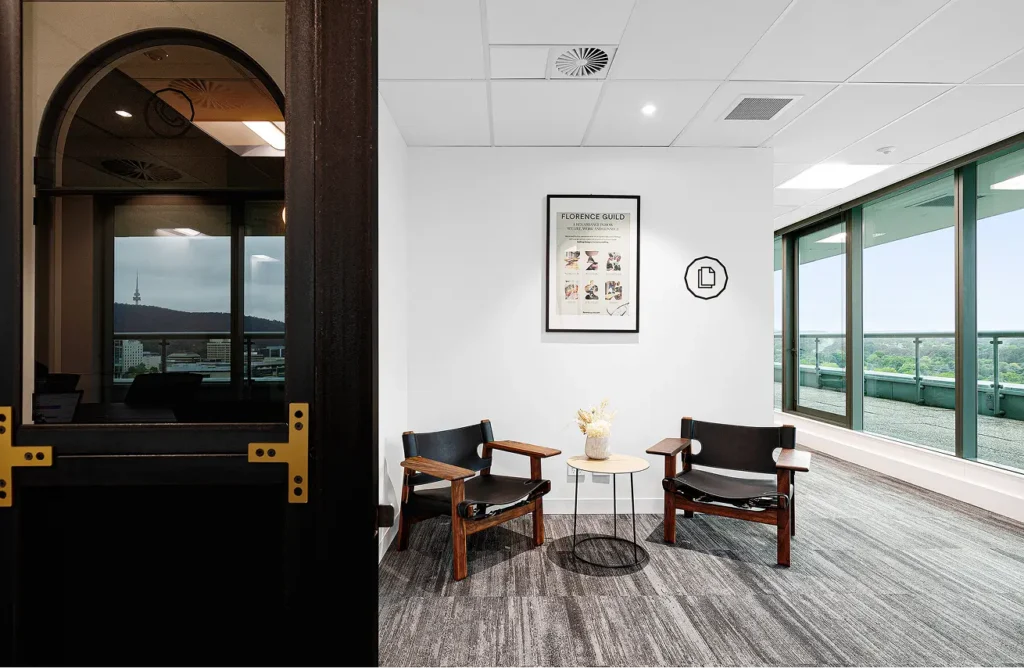 Two dark wood and black leather armchairs flank a small round table with flowers inside a modern, carpeted office room featuring large floor-to-ceiling windows. A framed poster on the wall reads: "FLORENCE GUILD A RENAISSANCE IN HOW WE LIVE, WORK AND CONNECT WORKPLACE WELLNESS ENTERTAINMENT DINING CULTURE EVENTS florenceguild.com"