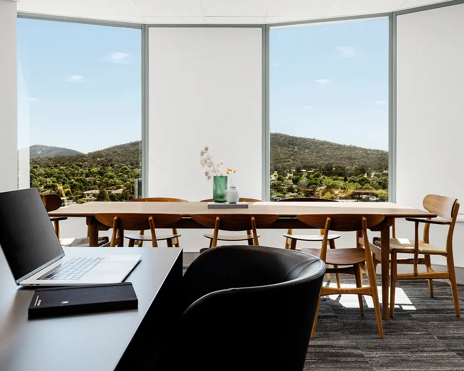 An open laptop rests on a desk facing a long wooden table decorated with vases on a book reading "CURATED," surrounded by large windows overlooking lush green hills.