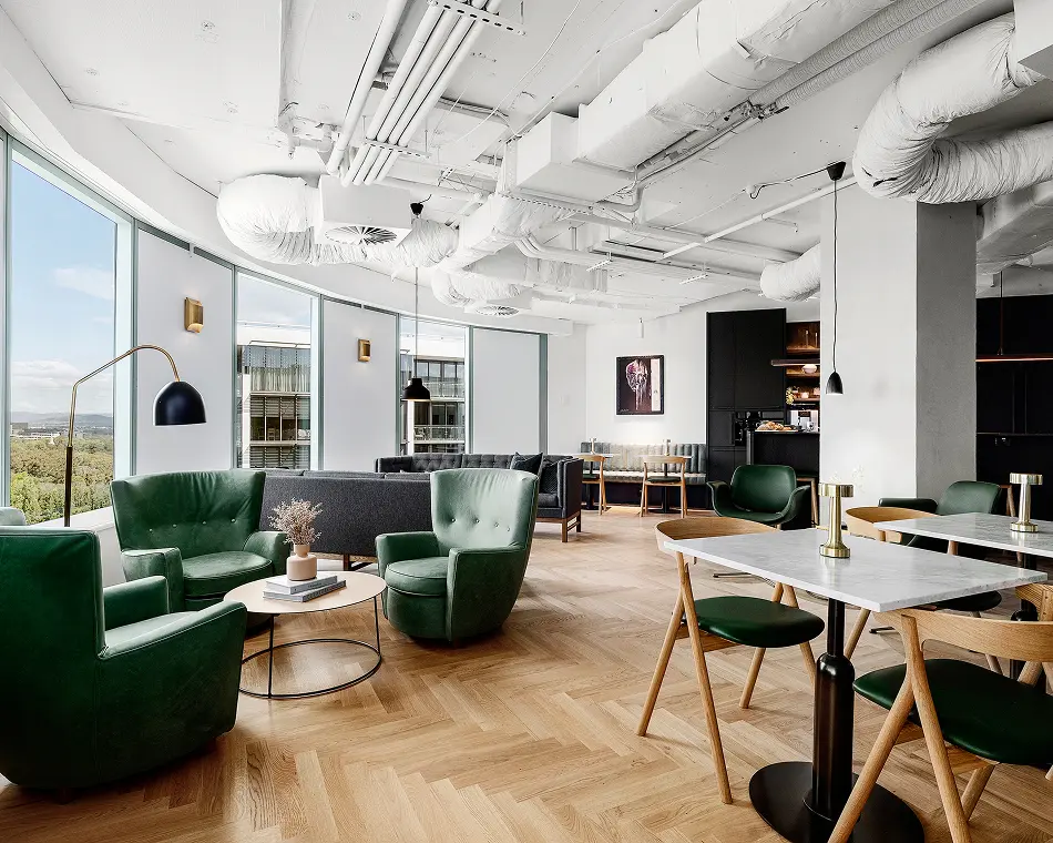 Green leather armchairs and marble-topped dining tables furnish a modern lounge area featuring light herringbone wood floors, large curved windows with city views, and exposed white ceiling ductwork.