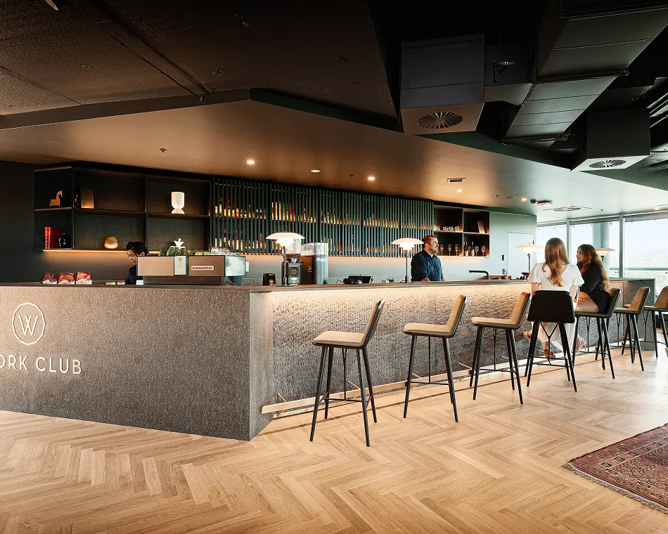 Patrons sit on high stools while staff serve behind a modern bar counter, situated in a lounge with light herringbone wood flooring and dark ceilings. Text on the counter reads: "W", "ORK CLUB".