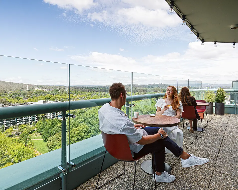 A man and woman sit enjoying drinks at a small table on a high-rise rooftop balcony, overlooking a lush green city park under a partly cloudy sky.