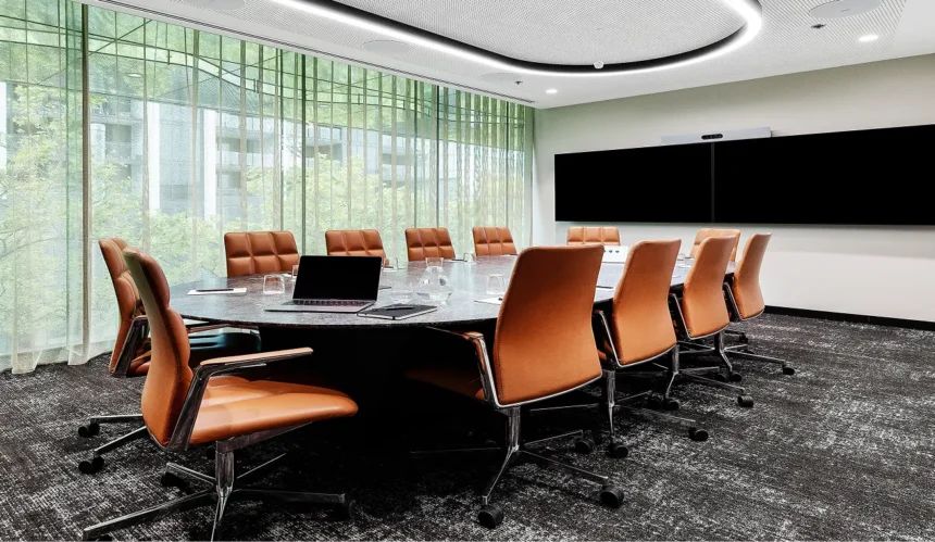 An oval dark stone conference table surrounded by brown leather office chairs sits prepared with laptops and water glasses inside a modern boardroom featuring large sheer-curtained windows.