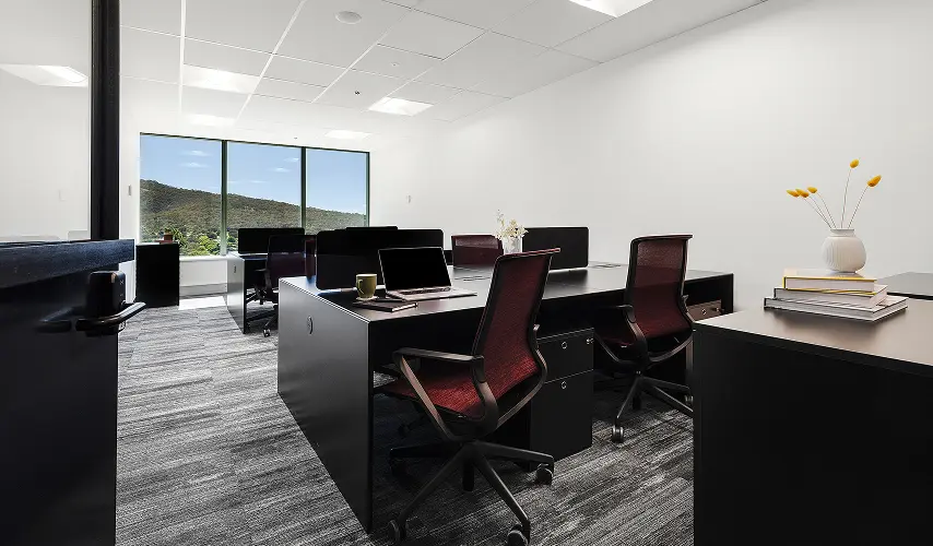 Black office workstations paired with dark red mesh chairs sit inside a bright, carpeted room featuring white walls and a large window overlooking a lush mountain landscape.