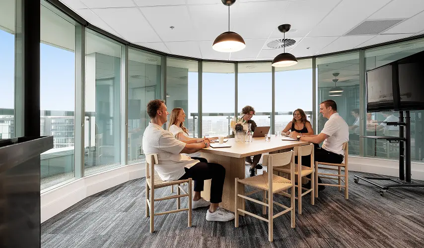 Five people sit around a light wooden conference table, conversing during a meeting inside a modern, glass-walled office room surrounded by panoramic city views and hanging dome lights.