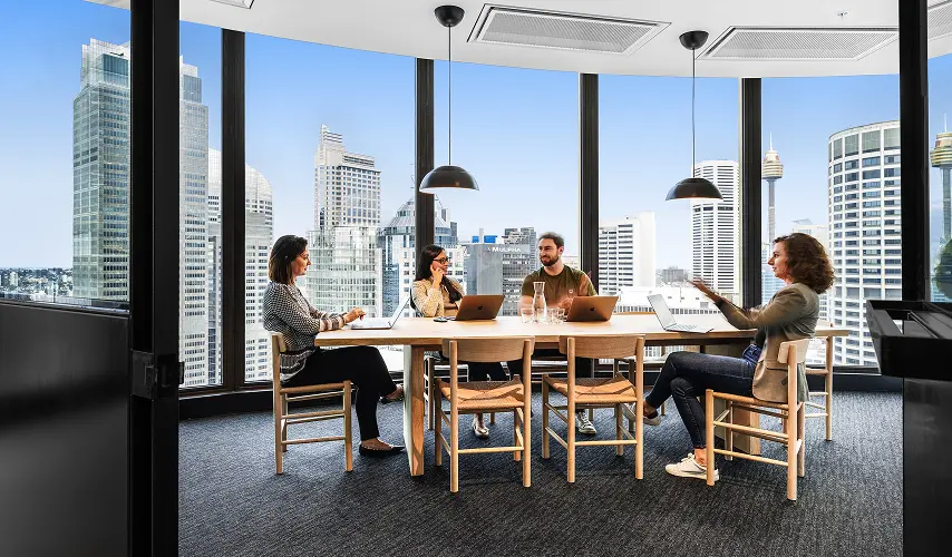 Four professionals sit at a wooden conference table with laptops, conversing inside a high-rise office with floor-to-ceiling windows that overlook a city skyline with a building displaying the text "MULPHA".