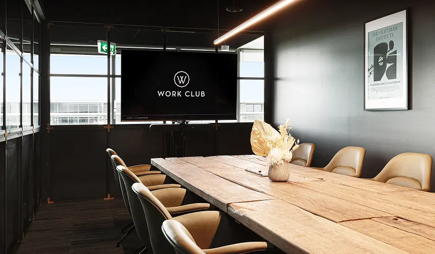 A rustic wooden conference table surrounded by beige chairs sits in a dark, modern boardroom, facing a large monitor displaying "W WORK CLUB". A wall poster reads "THE GOTTMAN INSTITUTE".