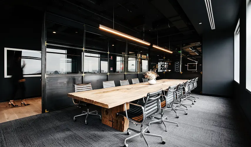 A long wooden conference table surrounded by modern black office chairs stands illuminated beneath linear pendant lights inside a dark boardroom featuring glass partition walls and grey carpet.
