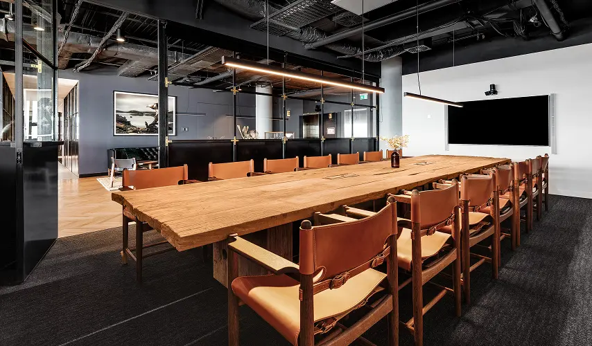 A long, rustic wooden conference table surrounded by leather chairs extends through a modern meeting room with dark carpet, glass partition walls, and exposed ceiling pipes.