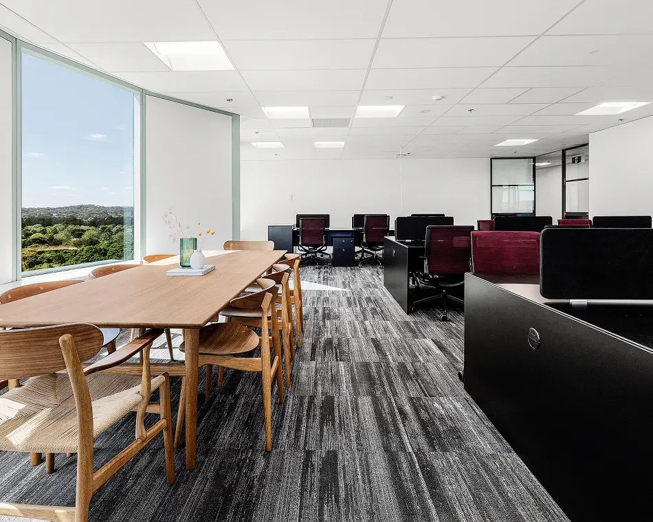 A long wooden conference table surrounded by chairs stands alongside black cubicle desks within a modern, bright office featuring large window views of lush greenery and patterned gray carpeting.