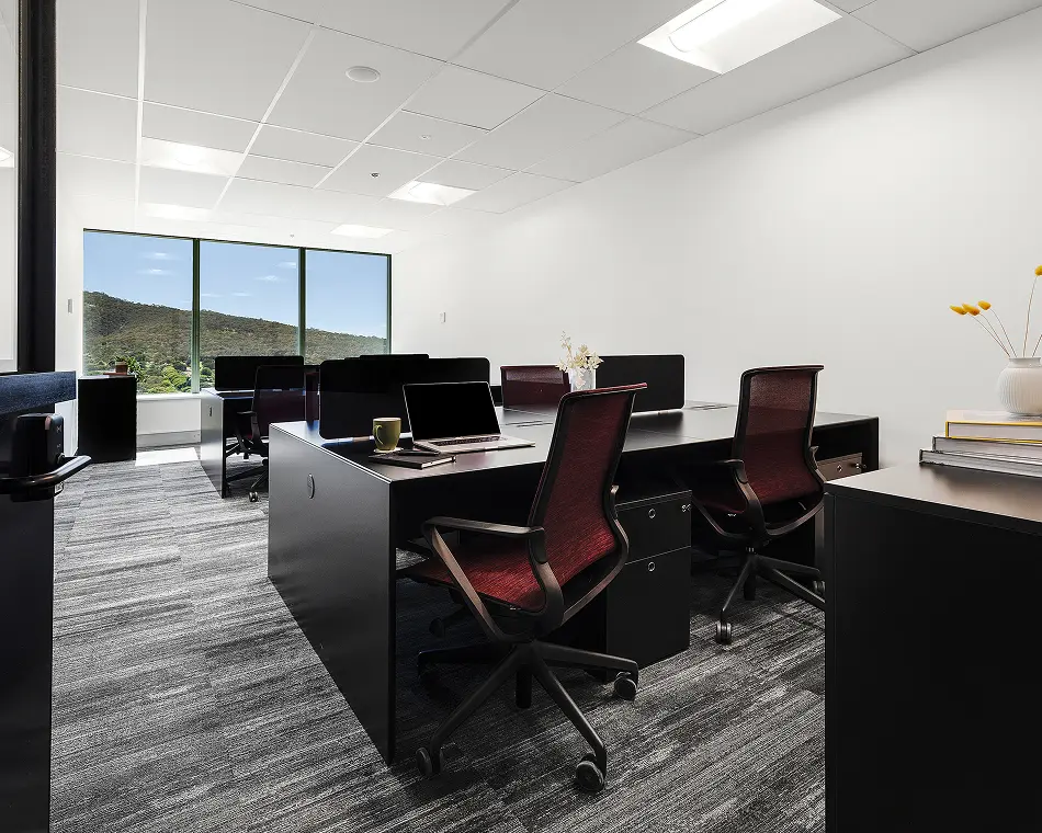 Black modern workstations with dark red mesh chairs sit inside a bright office space featuring grey carpet, white walls, and large windows displaying scenic mountain views.