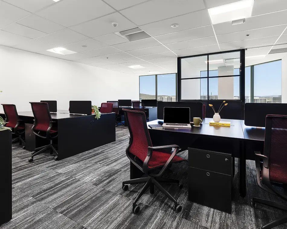 Black office desks paired with maroon mesh chairs stand arranged across a modern open-plan workspace featuring grey patterned carpet and large windows. A yellow book reads "Modernist Escapes".