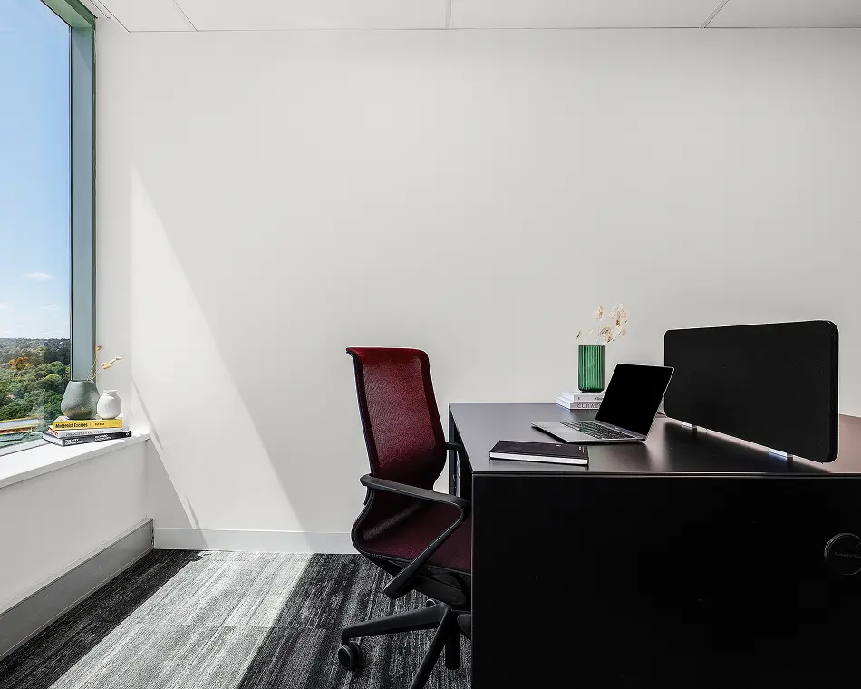 A burgundy mesh office chair sits beside a dark desk holding an open laptop and notebook, set in a minimalist white-walled office with a large window overlooking trees. Text on windowsill books reads: "Modernist Escapes", "PRINCIPLES OF STYLE", "THE CITY GARDENER", "RICHARD LANDSBERRY".