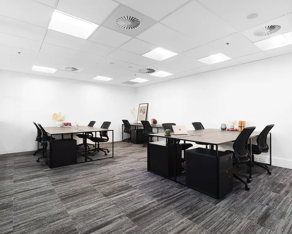 Sleek wooden office desks paired with black ergonomic chairs stand arranged in spacious clusters inside a modern, brightly lit room featuring plain white walls and patterned grey carpet.