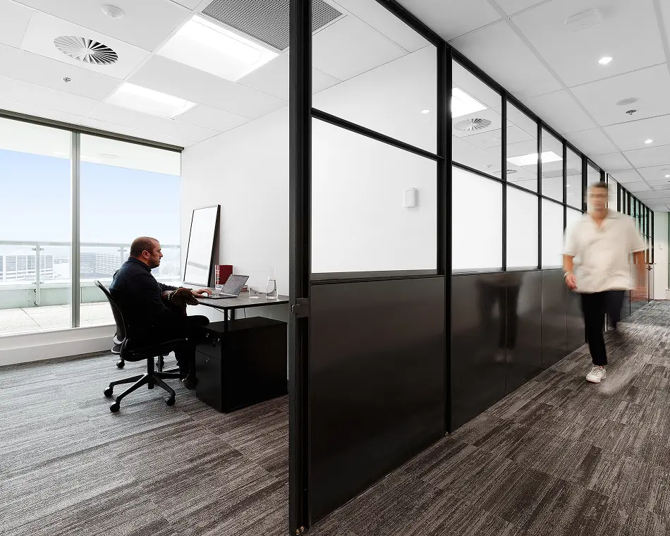 A man works on a laptop at a desk inside a glass-partitioned office room, while another blurred person walks past down the carpeted hallway.