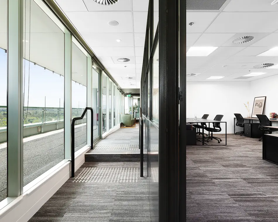 A carpeted office hallway lined with floor-to-ceiling windows extends toward a seating area, separated by a black-framed glass wall from an open workspace with desks and chairs.