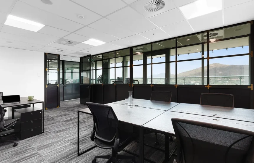 Black wooden desks and mesh office chairs sit in a modern workspace featuring patterned carpet, an acoustic tile ceiling, and grid-paneled glass walls overlooking distant outdoor hills. Text in image: "james. charley", "Humanscale".