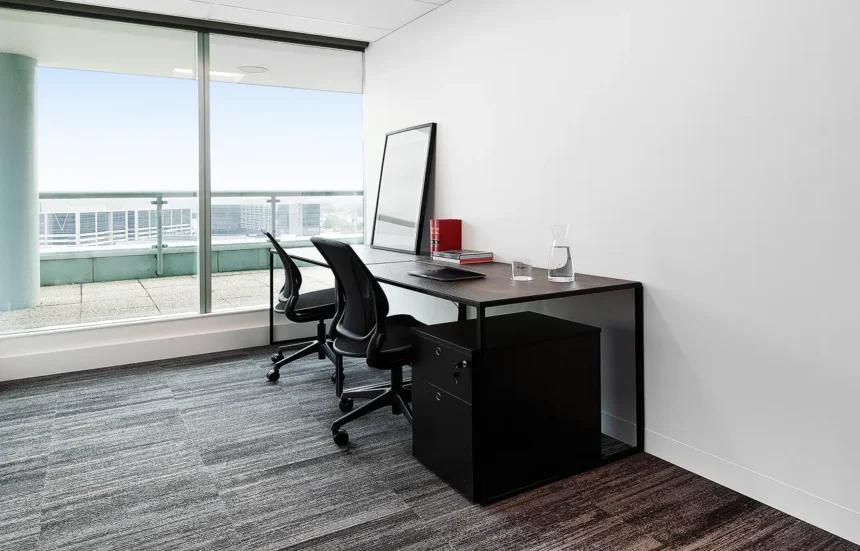 Two black office chairs sit at a long dark desk furnished with framed artwork and books, situated in a minimalist office room with white walls and floor-to-ceiling windows.