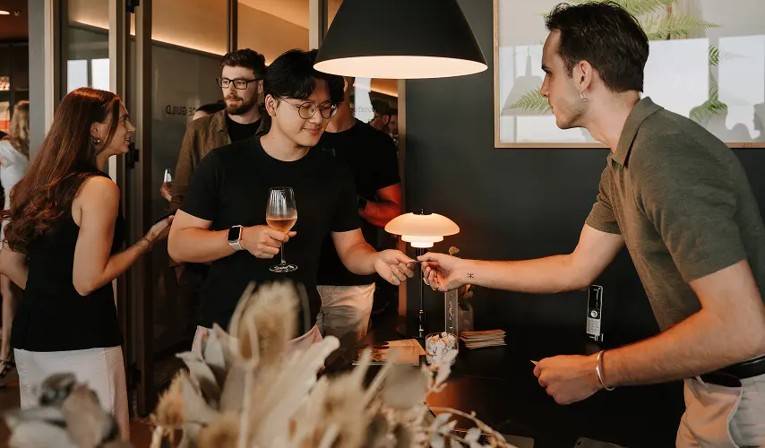 A man holding a wine glass hands a card to a host over a dark counter at an indoor gathering. Partially visible text on the glass door reads “ce guild”.