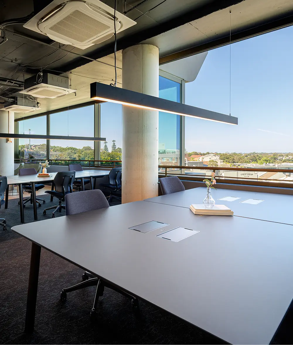 Sleek grey office desks and chairs sit arranged beneath suspended linear lights in a modern workspace, framed by floor-to-ceiling windows overlooking a sunny landscape featuring a building reading "CHIP SHOP".