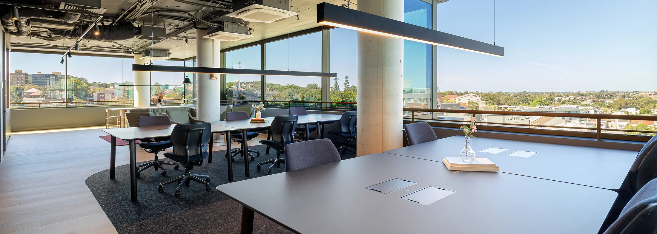 Sleek conference tables and ergonomic chairs furnish a modern open-plan office space, lined with floor-to-ceiling windows overlooking a sunny urban landscape with distant text reading "The Traditional CHIP SHOP".