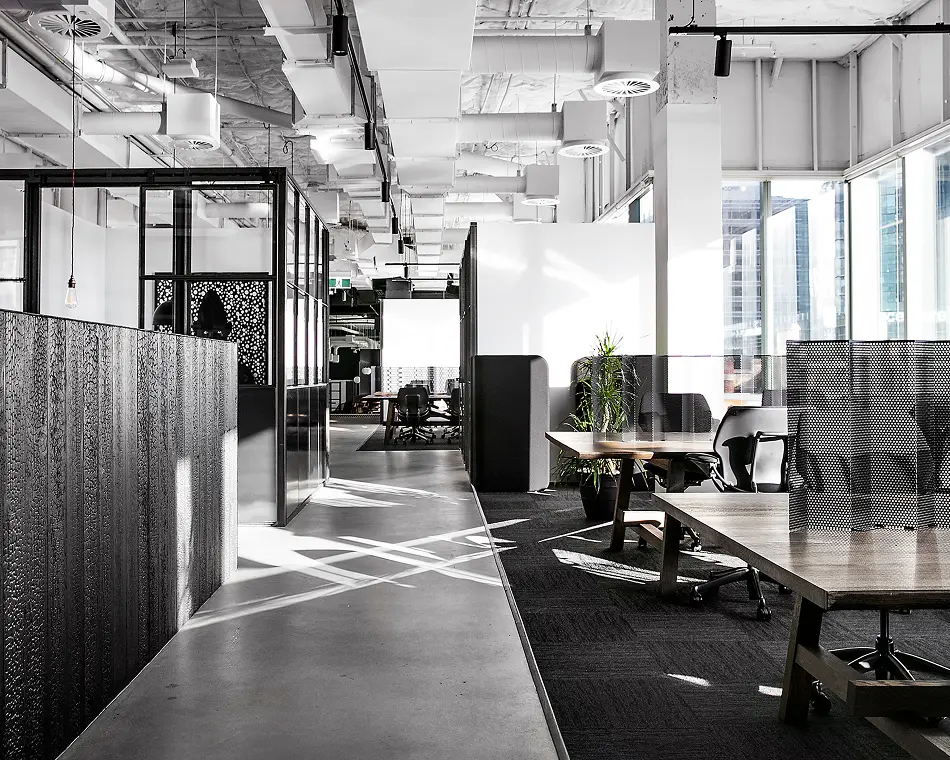 Wooden desks with black office chairs sit empty inside a modern, industrial office space, featuring exposed ceiling ductwork, a smooth concrete walkway, glass-walled enclosures, and large sunlit windows.