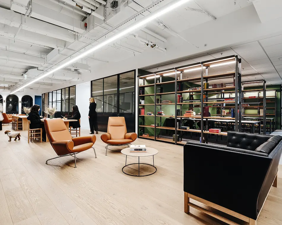 Two tan leather lounge chairs and a black armchair surround a low coffee table in a modern open-plan office space featuring light wood floors, exposed ceiling pipes, and metal shelving partitions.