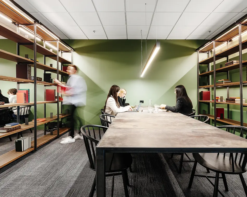 People work on laptops around a long wooden table while a motion-blurred man walks past tall illuminated bookshelves inside a modern study room featuring green walls.