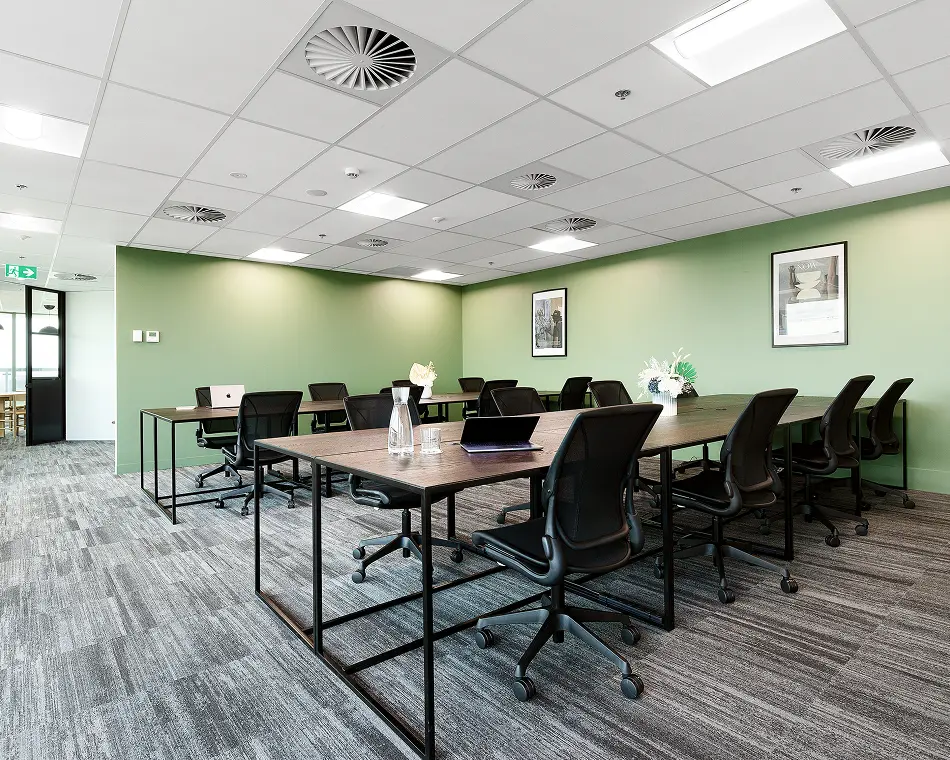 Long dark-wood desks lined with black ergonomic chairs stand arranged for shared work inside a modern office featuring light-green walls, patterned grey carpet, and a white drop ceiling.