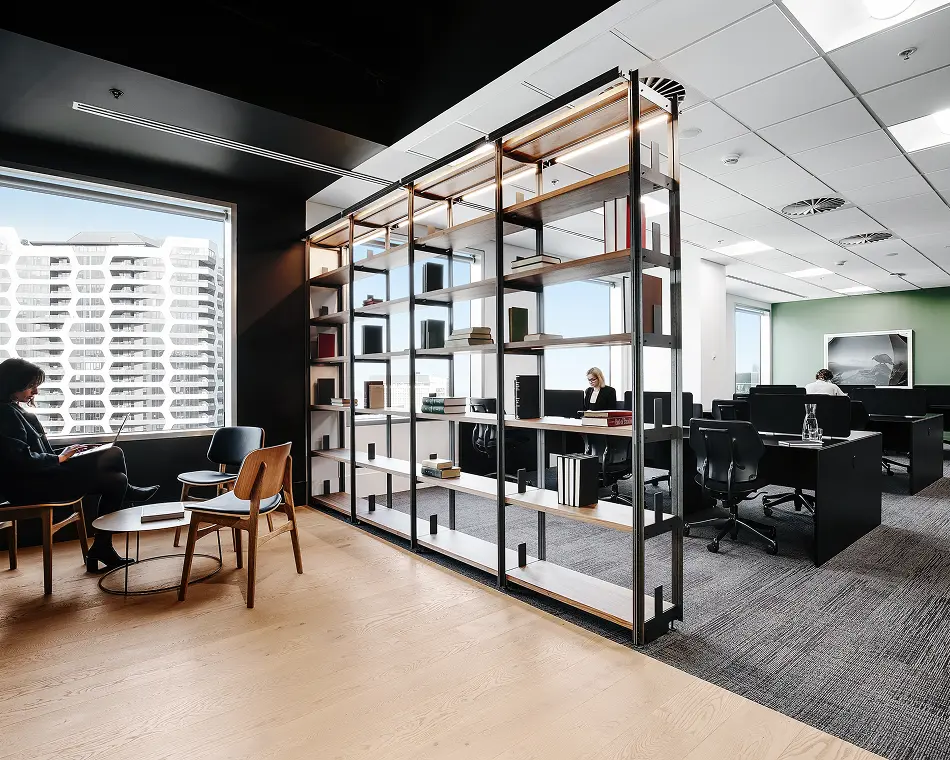 A tall metal and wood shelving unit divides a modern office space, separating a cozy seating lounge near large windows from an open desk workspace with working employees.