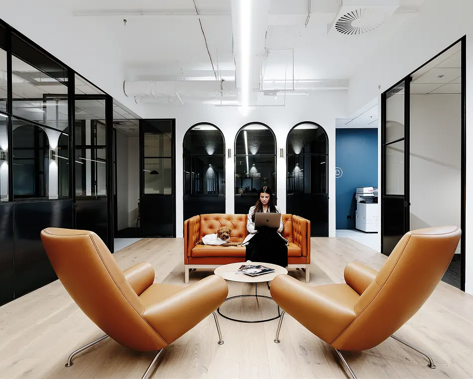 A woman sits on a brown leather sofa working on a laptop beside a small dog in a modern office lounge with two tan armchairs and black-framed glass walls.