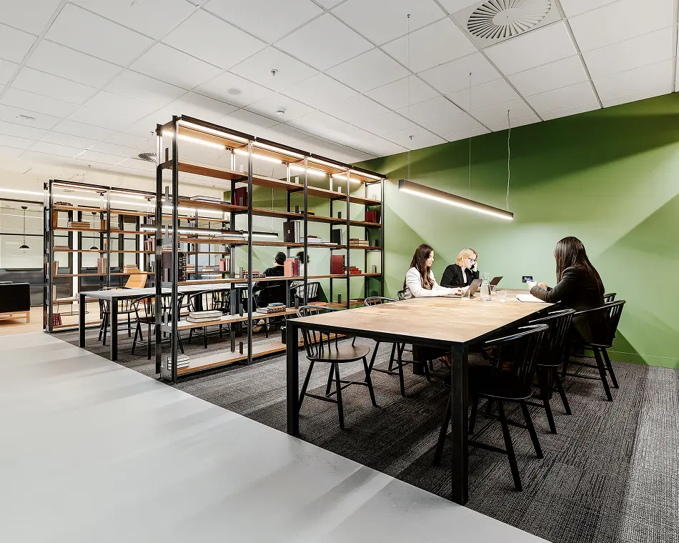 Office workers sit around a long wooden table working on laptops in a modern office space next to tall, open bookshelves and a vibrant green wall.