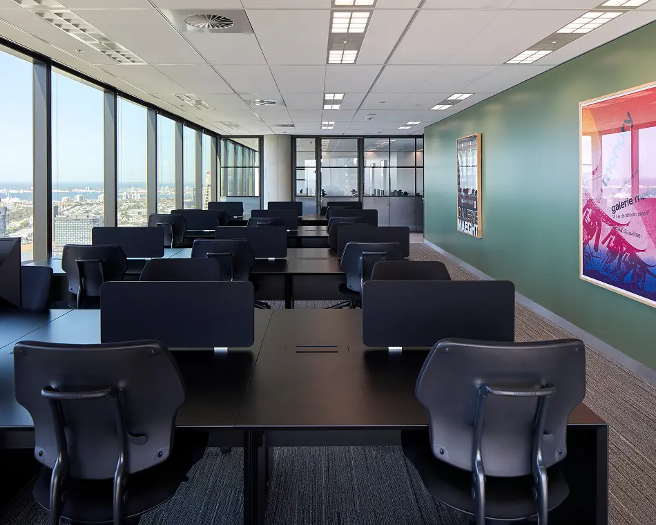 Rows of black office desks and ergonomic chairs fill a high-rise open-plan office, set beside floor-to-ceiling windows overlooking a cityscape and a green accent wall with framed art. Posters on the wall contain the text: "GALERIE MAEGHT" and "galerie maeght 13 rue de téhéran, paris 8e 30 avril 1981".