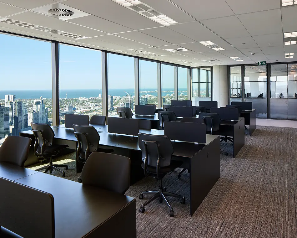 Black modern office desks paired with ergonomic chairs sit neatly arranged inside a high-rise workspace, surrounded by panoramic floor-to-ceiling windows overlooking a sunlit coastal city and ocean.
