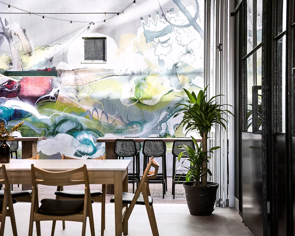 Modern wooden dining tables, chairs, and black bar stools sit arranged inside a bright patio space, situated beside a large colorful graffiti mural wall and a potted green houseplant.