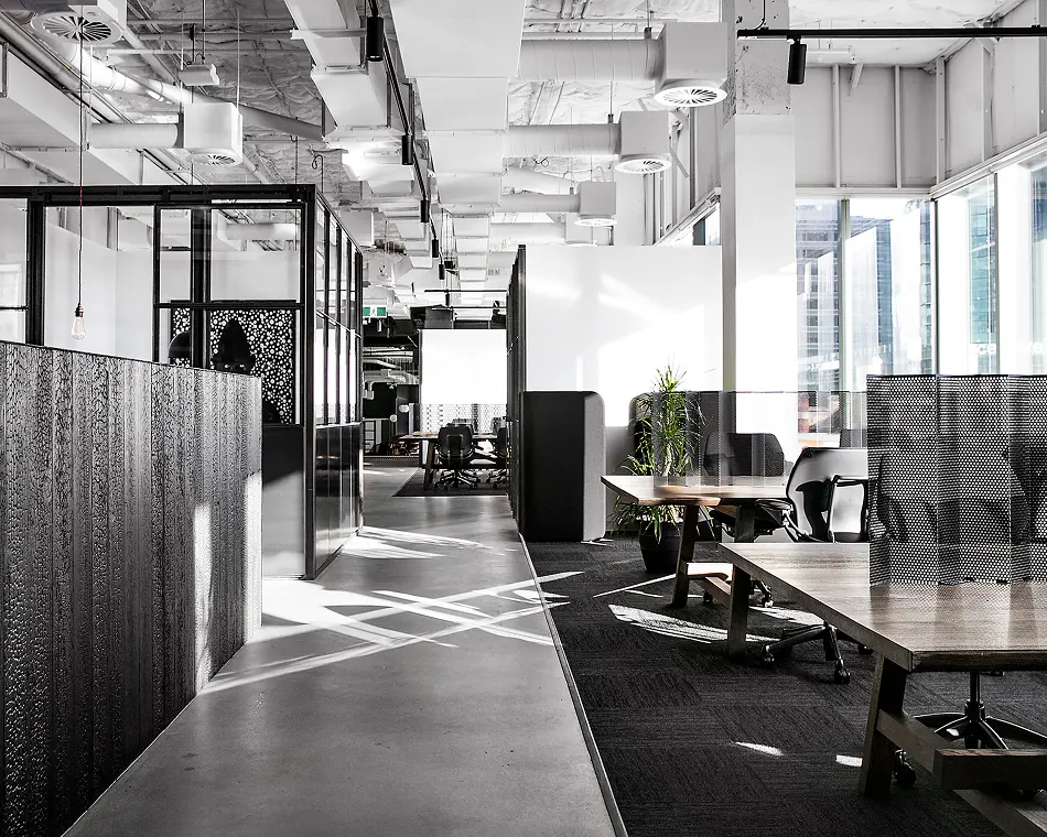 Empty wooden desks with ergonomic chairs stand inside a modern, sunlit office space featuring exposed white ceiling ducts, glass-framed meeting pods, and polished concrete flooring.