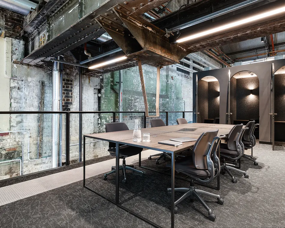 A wooden conference table surrounded by black ergonomic chairs sits inside a modern industrial office space featuring heavy exposed steel beams, distressed brick walls, and private work booths.