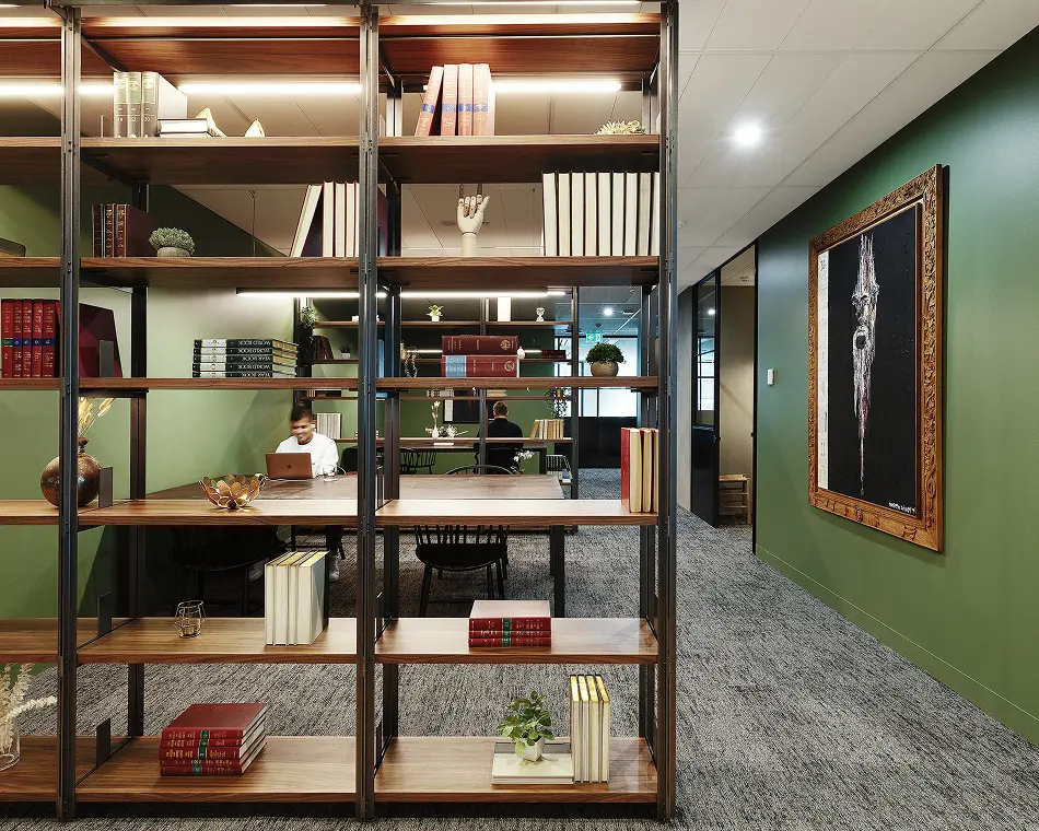 A large wooden and metal bookcase, filled with books and decor, acts as a room divider in a modern office, partially revealing people working at tables against green walls.
