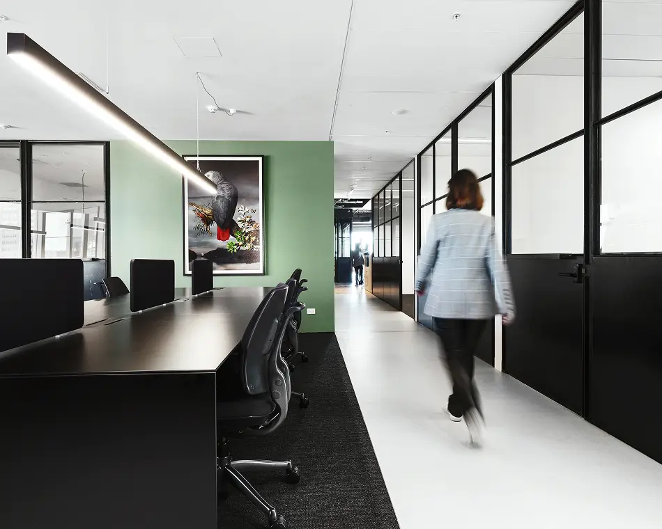 A person in a plaid blazer walks down a modern office hallway, set alongside black desks, ergonomic chairs, and glass-partitioned rooms with a green accent wall.