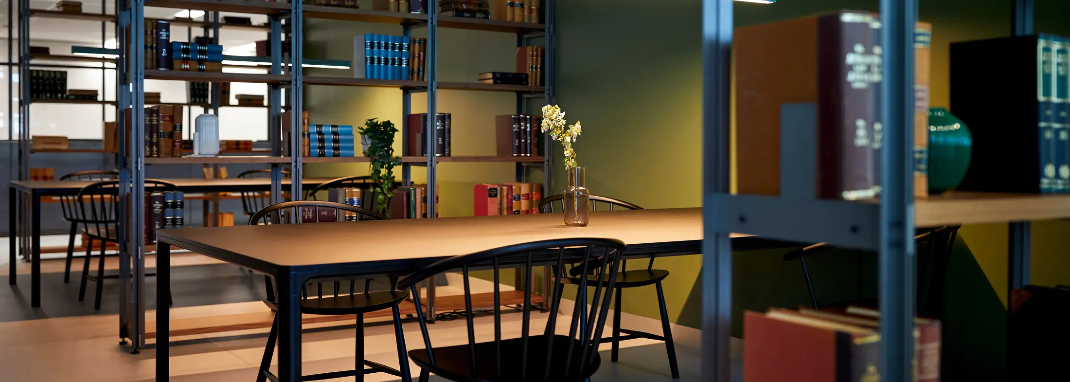 A long wooden study table surrounded by black chairs sits in a quiet, modern library, set against tall metal shelving units filled with books and a green wall.