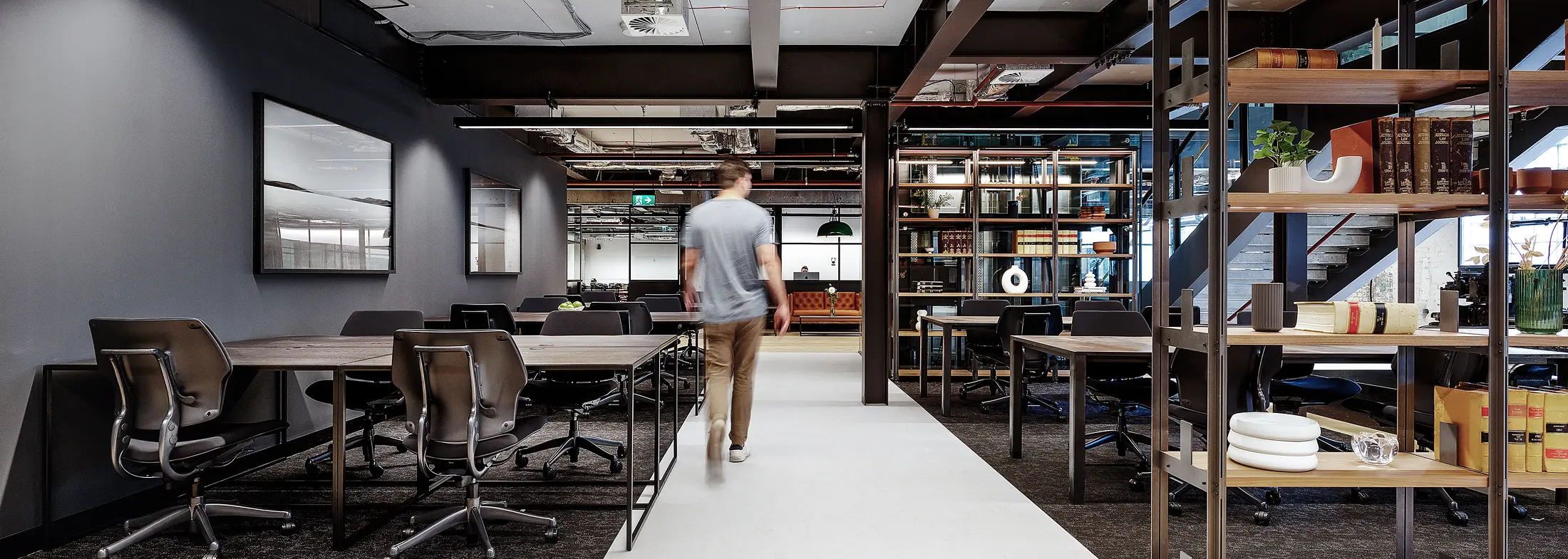 A man walks down a central white hallway within a modern, industrial office featuring black steel shelving, wooden desks with office chairs, dark grey walls, and exposed ceiling beams. Visible text on the book spines: "The Australian Law Journal", "Vol. 38 1964-65", "Vol. 39 1965-66", "Vol. 40 1966-67".