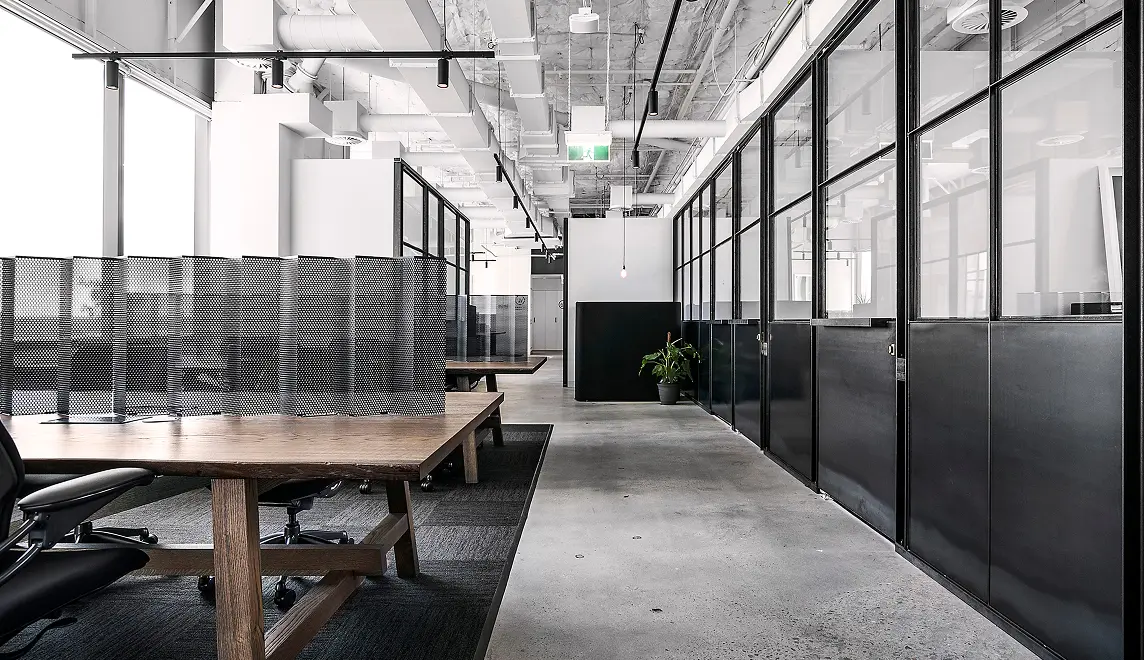 Wooden desks fitted with dark mesh privacy screens line an open corridor inside a modern office, featuring black-framed glass partitions, polished concrete floors, and exposed ceiling pipes.