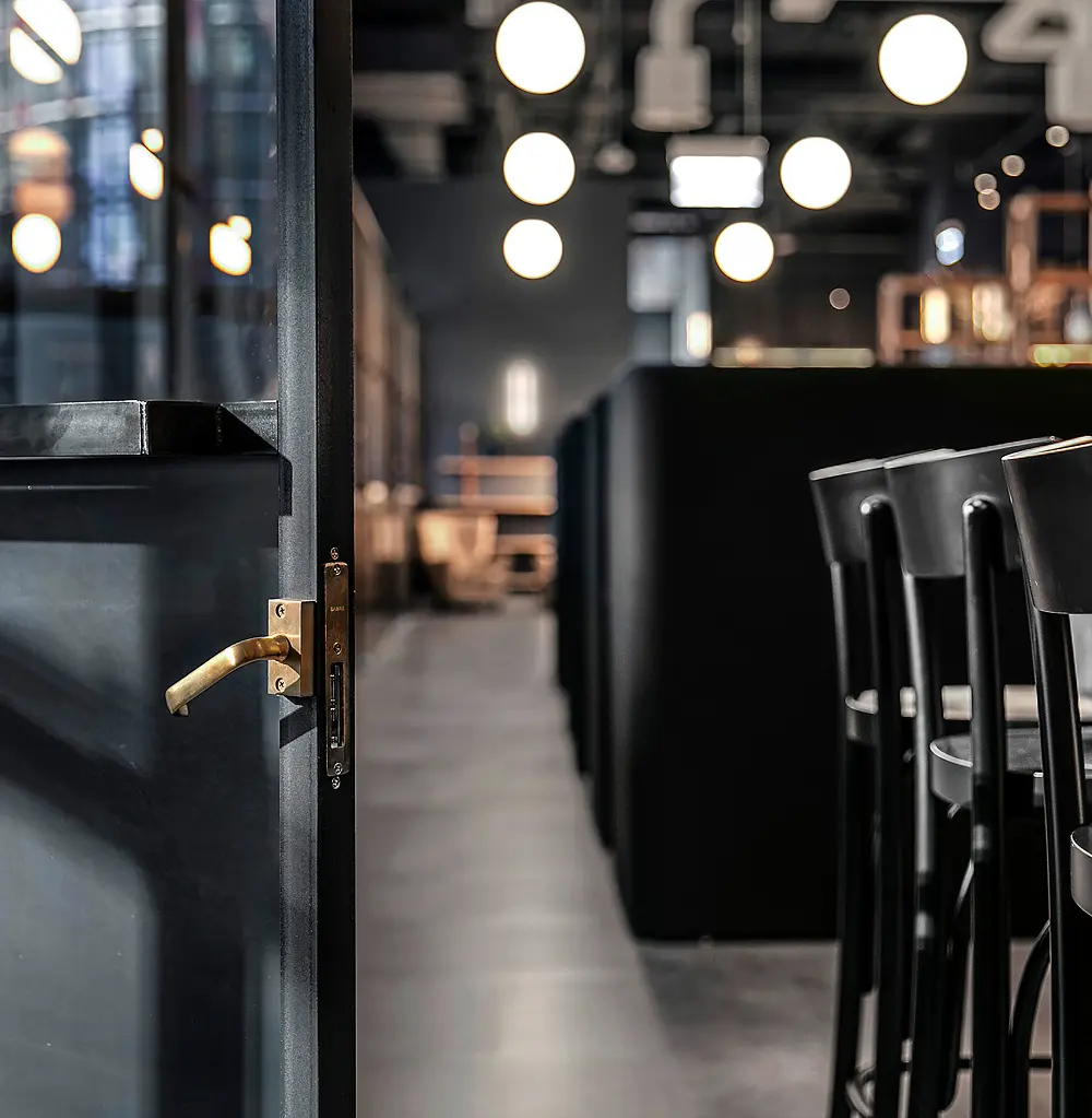 A dark grey door with a brass handle stands open, revealing a dimly lit, modern restaurant interior with glowing globe pendant lights, black bar stools, and dark booth seating.