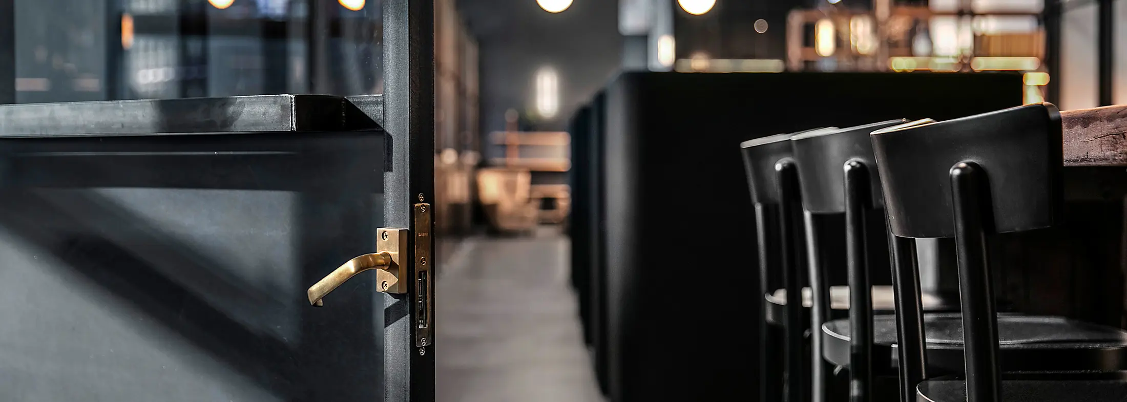 A sleek black door with a brass handle stands partially open, revealing a dimly lit, modern restaurant interior with a row of black bar stools under warm ambient lighting.
