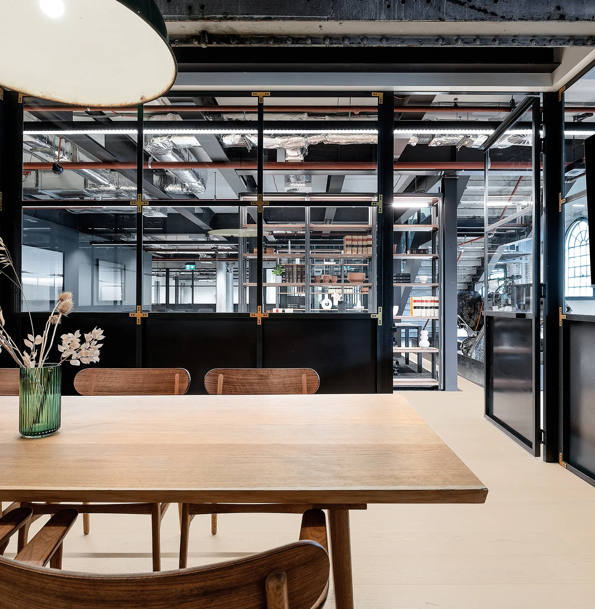 A light wooden table with chairs and a green flower vase stands in a modern office, positioned in front of black-framed glass partitions that reveal exposed industrial ceilings and adjacent workspaces.