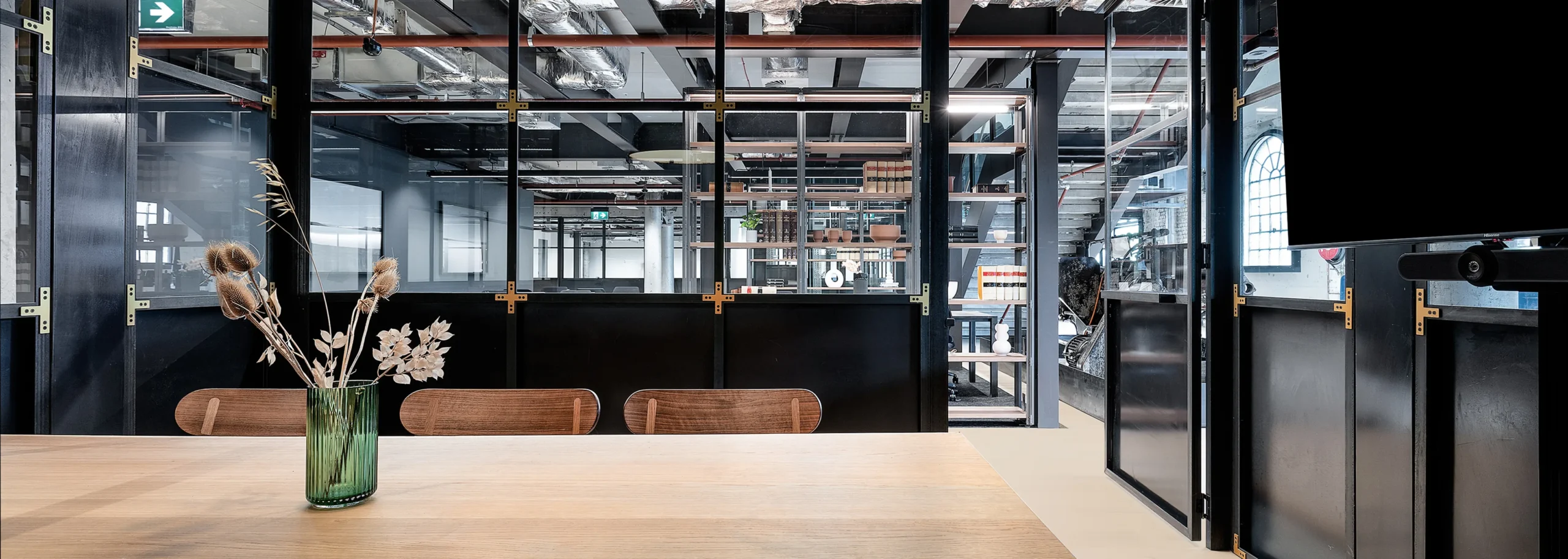 A light wooden table holding a green glass vase with dried flowers sits in front of wooden chairs, framed by black glass partitions in a modern, industrial-style office.