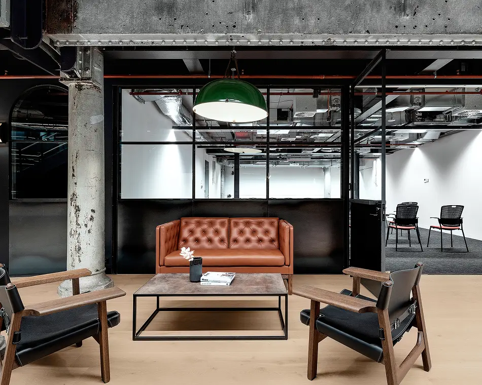 A brown leather tufted sofa, low coffee table, and two black armchairs form a seating area under a green dome pendant lamp inside a modern industrial office space.